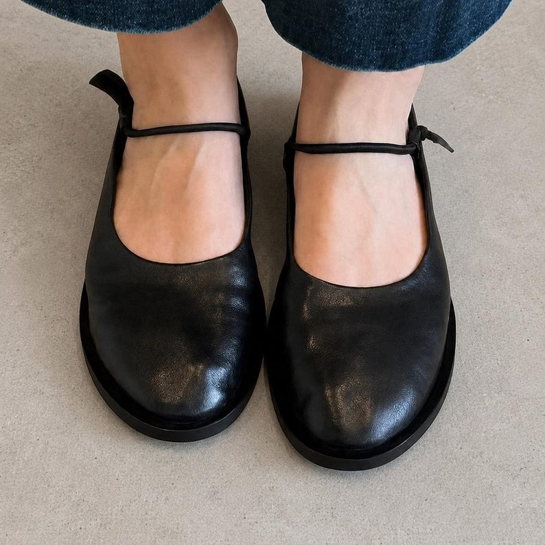 May include: A pair of black leather Mary Jane shoes featuring a rounded toe, a smooth finish, and a thin, adjustable strap that secures across the top of the foot with a small knot detail. The shoes have a flat sole and a low-cut opening.