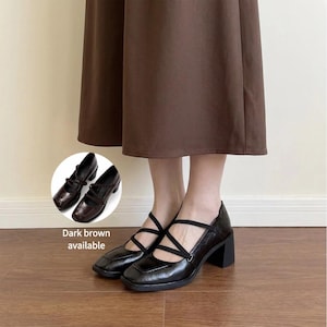 May include: A pair of dark brown block-heeled shoes with a square toe and two thin, crisscrossing elastic straps across the top. The shoes feature a textured finish and a sturdy heel. An inset shows the shoes from a different angle.