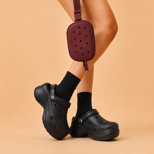 May include: Black platform clogs with a strap, paired with black socks. A burgundy fanny pack with a dotted pattern hangs from the leg. The background is a solid peach color.