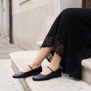 May include: A pair of black leather ballet flats featuring a thin strap across the instep and a low heel. The shoes are paired with a black lace skirt that falls to the mid-calf, showcasing a smooth finish on the footwear and intricate lace.