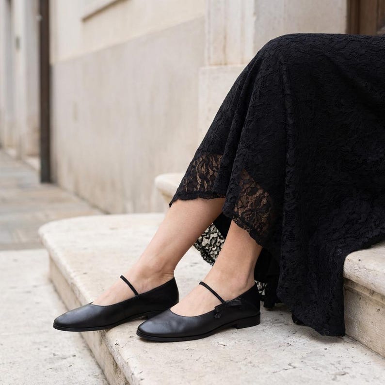 May include: A pair of black leather ballet flats featuring a thin strap across the instep and a low heel. The shoes are paired with a black lace skirt that falls to the mid-calf, showcasing a smooth finish on the footwear and intricate lace.