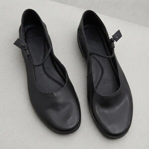 May include: A pair of black flat shoes featuring a rounded toe and a thin strap across the instep. The shoes have a smooth finish, a low-cut opening, and a simple design with a small knot detail on the side of each strap, providing a clean look.