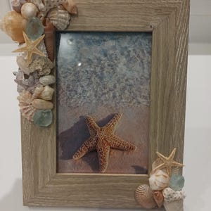 May include: A rectangular picture frame with a light brown wood grain finish. The frame is decorated with an assortment of seashells, small starfish, and pieces of sea glass attached to the top left and bottom right corners of the frame.