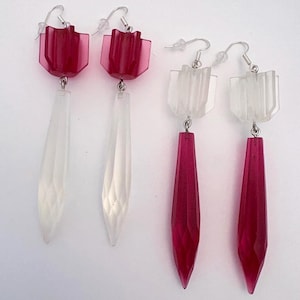 May include: Two pairs of drop earrings featuring faceted, translucent, elongated pendants suspended from geometric, finned tops. One pair has deep magenta tops with clear pendants, while the other pair has clear tops with deep magenta pendants.