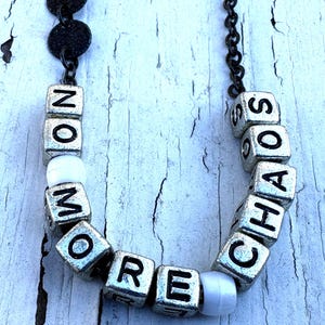 May include: A necklace featuring a dark metal chain with silver-toned cube beads. The beads are embossed with black letters spelling out the phrase NO MORE CHAOS. The design includes small white spherical beads interspersed between the lettered cubes.