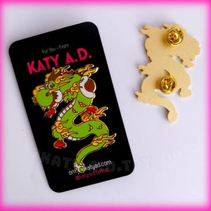 May include: Green and gold enamel pin featuring a cartoon dragon with flames and clouds. The pin is on a black card with the text "For You - From KATY A.D." and a website address.