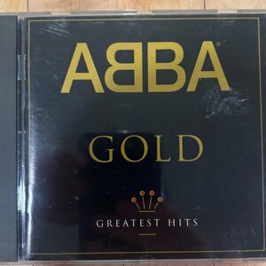 Könnte beinhalten: Eine schwarze CD-Hülle mit dem Cover des Albums ABBA Gold Greatest Hits. Das Front-Inlay zeigt den Bandnamen mit einem gespiegelten ersten B in goldener Schrift, das Wort GOLD in Gold und den Text GREATEST HITS unter einem kleinen Kronensymbol.