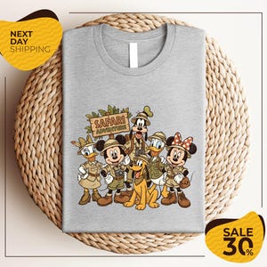 A gray t-shirt with a printed design of Disney characters dressed in safari attire, including Minnie Mouse, Goofy, Donald Duck, and Mickey Mouse, along with a sign that says "Safari Adventure".