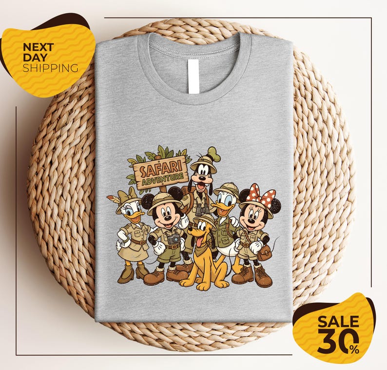 A gray t-shirt with a printed design of Disney characters dressed in safari attire, including Minnie Mouse, Goofy, Donald Duck, and Mickey Mouse, along with a sign that says "Safari Adventure".