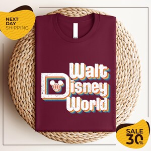 a maroon t-shirt with the text "Walt Disney World" printed on it, along with a Mickey Mouse logo. The t-shirt is displayed on a woven basket, and there are two yellow price tags in the image.