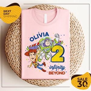A pink t-shirt with a cartoon image of Buzz Lightyear and Woody from Toy Story, along with the text "Olivia 2 Infinity Beyond". The t-shirt is displayed on a woven basket, and there are yellow price tags with text indicating a sale of 30% off.