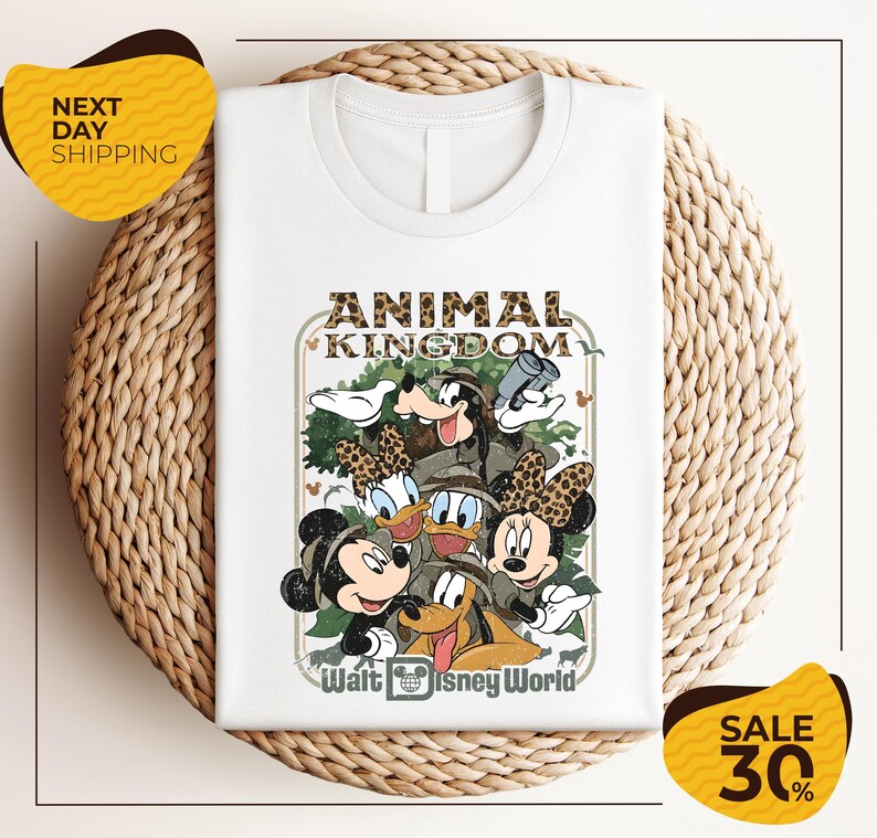 A white t-shirt with a Disney-themed graphic design featuring Minnie Mouse, Donald Duck, and Goofy, along with the text "Animal Kingdom" and " Walt Disney World". The t-shirt is displayed on a woven placemat, and there are yellow sale signs indicating a 30% discount.