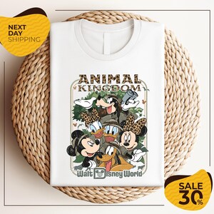 A white t-shirt with a Disney-themed graphic design featuring Minnie Mouse, Donald Duck, and Goofy, along with the text "Animal Kingdom" and " Walt Disney World". The t-shirt is displayed on a woven placemat, and there are yellow sale signs indicating a 30% discount.