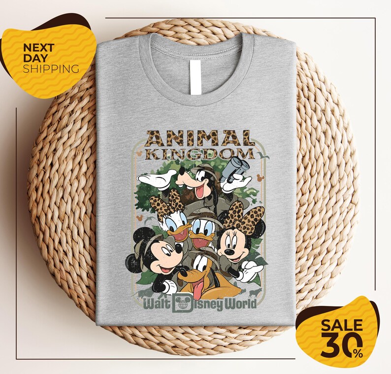 A gray t-shirt with a graphic design featuring Disney characters Mickey Mouse, Minnie Mouse, Donald Duck, and Goofy, along with the text "Animal Kingdom" and " Walt Disney World". The t-shirt is displayed on a woven placemat, and there are yellow and black sale signs indicating a 30% discount.