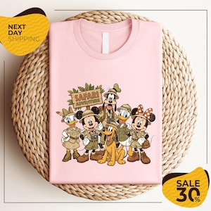 A pink t-shirt with a cartoon illustration of Disney characters dressed in safari attire, including Minnie Mouse, Goofy, Donald Duck, and Pluto, along with a sign that says "SAFARI ADVENTURE". The t-shirt is displayed on a woven placemat, and there are yellow sale signs indicating a 30% discount.