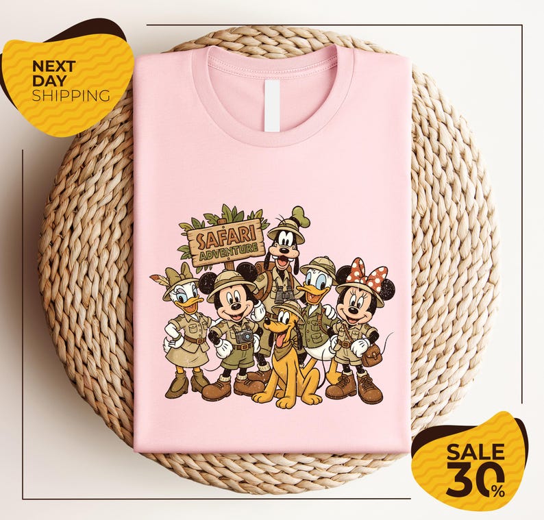 A pink t-shirt with a cartoon illustration of Disney characters dressed in safari attire, including Minnie Mouse, Goofy, Donald Duck, and Pluto, along with a sign that says "SAFARI ADVENTURE". The t-shirt is displayed on a woven placemat, and there are yellow sale signs indicating a 30% discount.