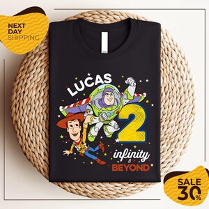 A black t-shirt with a colorful graphic design featuring characters from the Toy Story franchise, including Buzz Lightyear and Woody, along with the text "Lucas 2 infinity & beyond".