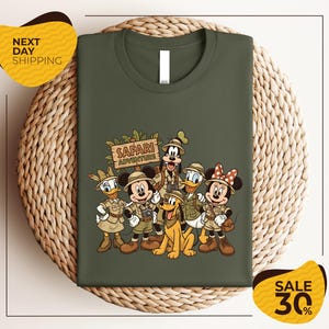 A green t-shirt with a printed illustration of Disney characters dressed in safari attire, including Minnie Mouse, Donald Duck, Goofy, and Mickey Mouse, along with a sign that says "SAFARI ADVENTURE".
