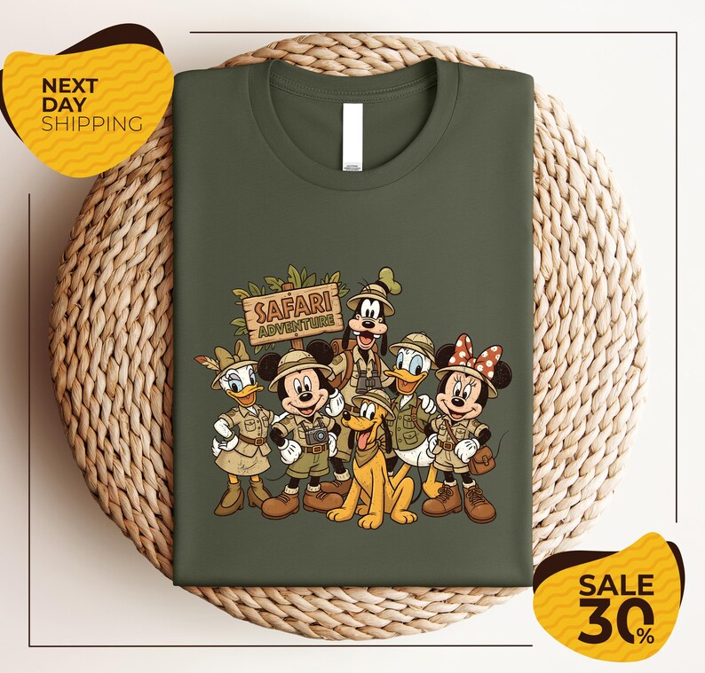 A green t-shirt with a printed illustration of Disney characters dressed in safari attire, including Minnie Mouse, Donald Duck, Goofy, and Mickey Mouse, along with a sign that says "SAFARI ADVENTURE".