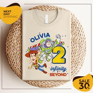 a t-shirt with a cartoon character from the Toy Story franchise, specifically Woody, and the text "Olivia 2 Infinity Beyond". The t-shirt is displayed on a woven basket, and there are two yellow price tags in the image.
