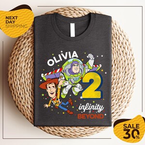 a gray t-shirt with a design of two characters from the Toy Story franchise, specifically Buzz Lightyear and Woody, along with the text "Olivia 2 infinity & beyond". The t-shirt is displayed on a woven placemat, and there are two yellow price tags in the image, one on the left and one on the right, indicating a sale or discount.