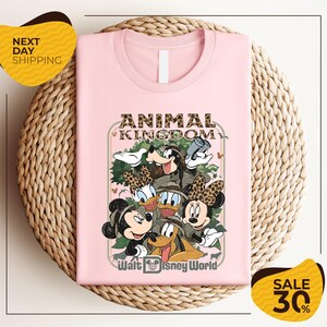 A pink t-shirt with a Disney-themed graphic design, featuring characters from the Disney movie "Animal Kingdom" and the text "Walt Disney World". The t-shirt is displayed on a woven basket, and there are two yellow sale signs indicating a 30% discount.