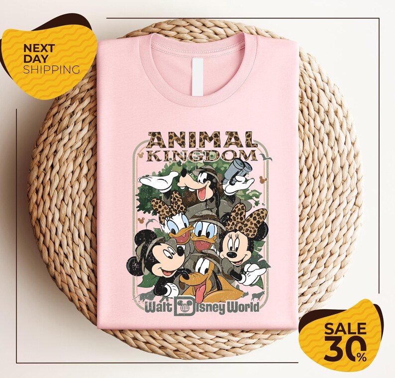 A pink t-shirt with a Disney-themed graphic design, featuring characters from the Disney movie "Animal Kingdom" and the text "Walt Disney World". The t-shirt is displayed on a woven basket, and there are two yellow sale signs indicating a 30% discount.