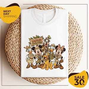 A white t-shirt with a printed illustration of Disney characters dressed in safari attire, including Minnie Mouse, Goofy, Donald Duck, and Mickey Mouse, along with a sign that says "SAFARI ADVENTURE".