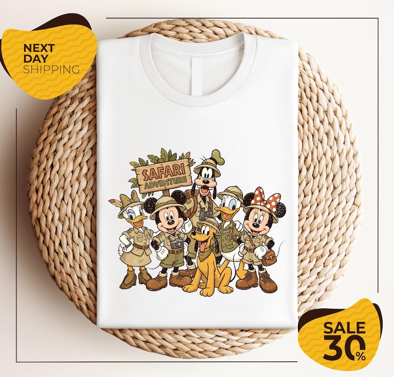 A white t-shirt with a printed illustration of Disney characters dressed in safari attire, including Minnie Mouse, Goofy, Donald Duck, and Mickey Mouse, along with a sign that says "SAFARI ADVENTURE".