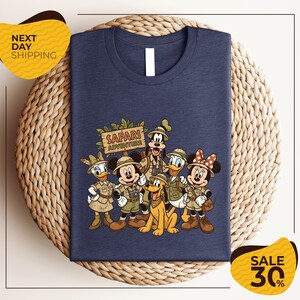 A blue t-shirt with a printed design of Disney characters dressed in safari attire, including Mickey Mouse, Minnie Mouse, Donald Duck, Goofy, and Pluto, along with a safari adventure sign. The t-shirt is displayed on a woven placemat, and there are yellow price tags indicating a sale of 30% off.