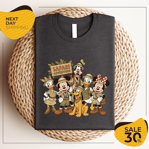 A t-shirt featuring a cartoon illustration of Disney characters dressed in safari attire, including Mickey Mouse, Minnie Mouse, Donald Duck, Goofy, and Pluto, along with a sign that says "Safari Adventure".