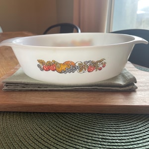 May include: White oval casserole dish with a fruit design featuring strawberries, grapes, and oranges. The dish has two handles and is resting on a wooden cutting board and a green placemat. A vintage kitchenware item.