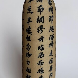 May include: A cylindrical ceramic bottle with a matte beige finish and a black neck. The body features vertical columns of black Chinese calligraphy characters. The base is decorated with a black geometric border pattern on a light background.