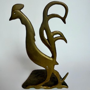 May include: A brass bookend shaped like a stylized rooster in profile. The metal figure features a smooth, matte gold-toned finish with cut-out sections defining the tail feathers and neck. It stands on a flat, dark-toned base plate.