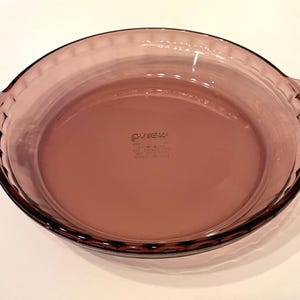 May include: A vintage Pyrex pie dish in a soft, translucent purple hue. The round dish features a fluted rim and integrated handles. The Pyrex logo is visible on the base. A classic kitchenware item.