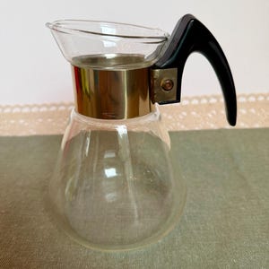 Peut inclure: Une carafe en verre transparent dotée d'un corps large et conique avec un bord évasé. Le col est entouré d'une bande métallique dorée polie, reliée à une poignée incurvée en plastique noir fixée par une vis visible sur le côté.
