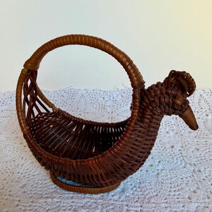 May include: A brown woven wicker basket shaped like a bird with a rounded handle. The basket features a detailed head with a pointed beak and a crest, constructed from thin, flexible reeds woven into a sturdy, open-topped container structure.
