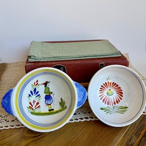 May include: Two ceramic bowls with hand-painted designs. The left bowl features a figure in a green jacket and blue striped pants with blue handles. The right bowl displays a large red flower with green leaves and blue circular rim accents.