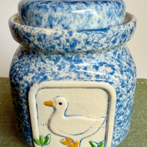 May include: A ceramic canister with a blue and white sponge-painted finish and a matching lid. The front features a square, cream-colored relief panel depicting a white duck with an orange beak and feet, accented by small green grass blades.