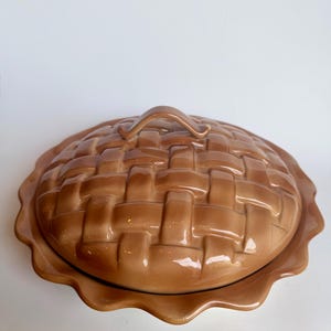 May include: A round, light brown ceramic pie dish with a matching lid. The lid features a raised, embossed lattice pattern resembling a woven pie crust, complete with a central handle. The dish has a decorative, scalloped, ruffled edge.