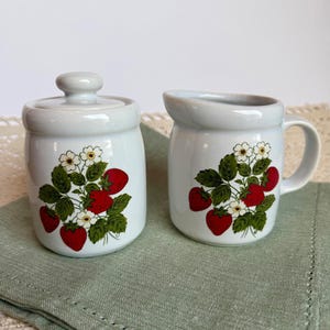 May include: A white ceramic sugar bowl with a lid and a matching creamer jug, both decorated with a botanical print of red strawberries, green leaves, and small white flowers on the front surface of each piece.