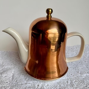 May include: A teapot featuring a polished copper-coloured metallic body with a smooth, reflective finish. It includes a cream-coloured ceramic spout, handle, and base, topped with a small, round, gold-toned metal knob on the lid.