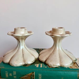 May include: A pair of off-white ceramic candle holders with a fluted, petal-like base and a smooth, cylindrical top opening. The surface features a subtle, weathered brown patina along the grooves and edges, giving a textured appearance.