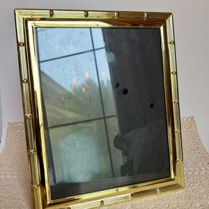 May include: A rectangular picture frame with a polished gold-tone finish and a decorative bamboo-style molded border. The frame features a clear glass front and a dark interior, reflecting light and surrounding objects on its reflective surface.