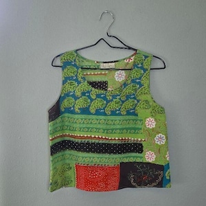 May include: A sleeveless tank top featuring a patchwork design with various patterns in shades of green, blue, black, and red. The garment includes floral motifs, dotted textures, and paisley-like shapes, finished with a round neckline.