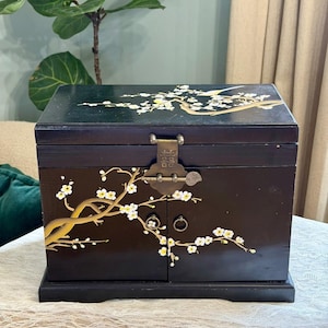 May include: A black wooden jewelry box featuring a hand-painted white and gold plum blossom design. The box includes a hinged lid, two front doors with small circular metal pulls, and a central brass latch engraved with a double happiness symbol.