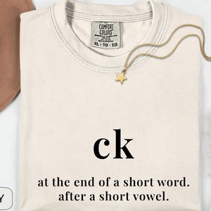 May include: Ivory-colored t-shirt with the letters "ck" in black, and the text "at the end of a short word. after a short vowel." A gold star pendant necklace and a brown hat are also visible.