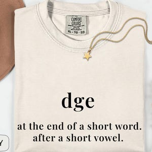 May include: Ivory-colored t-shirt with the word "dge" printed in black, along with the phrase "at the end of a short word. after a short vowel." A gold star necklace and a brown hat are also visible.