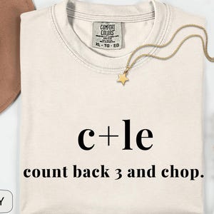 May include: Cream-coloured t-shirt with the text "c+le count back 3 and chop." A gold star pendant necklace rests on the shirt. The shirt has a Comfort Colors tag. A brown hat and flowers are in the background.
