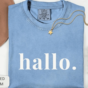 May include: A light blue crew neck t-shirt with the word "hallo." printed in white on the front. The shirt features a Comfort Colors brand label at the inner neckline, a gold-toned chain necklace with a star pendant, a brown hat, and dried flowers.